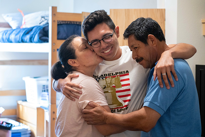 A student closed their eye and smiles as their parents hug them in their dorm room.