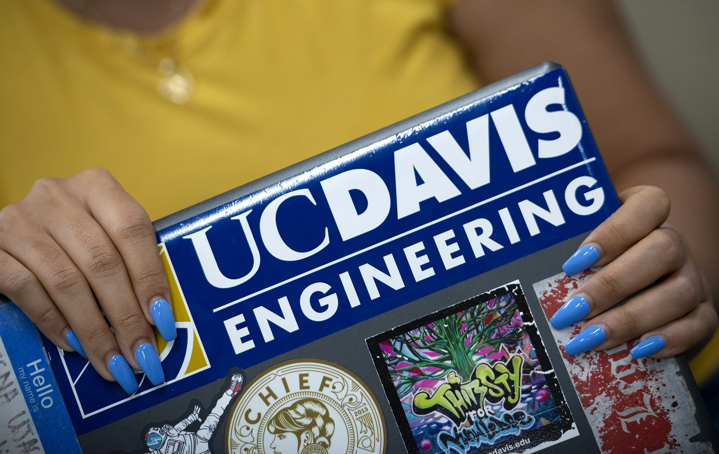 Closeup view of a person's hands while blue manicured nails holding a laptop computer covered in a variety of stickers. The largest sticker reads "UC Davis Engineering."