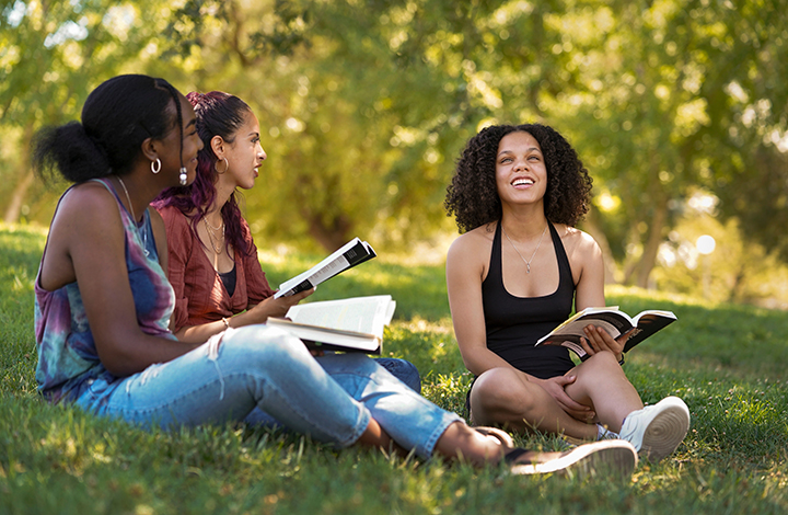 Three students holding books sit on the lawn talking to one another.