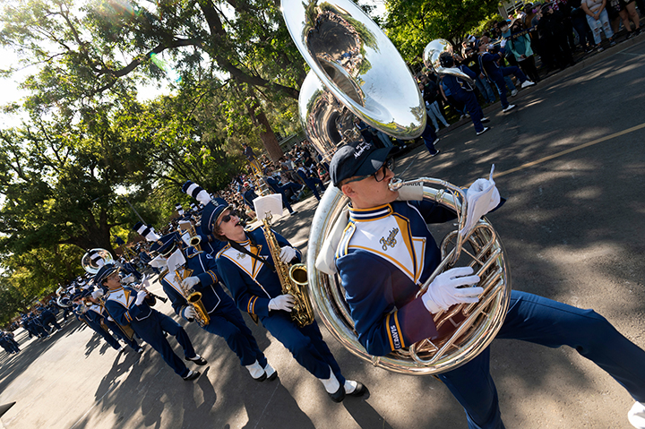 A student playing a tuba leads a line of marching band members as they play their instruments and march in a parade.
