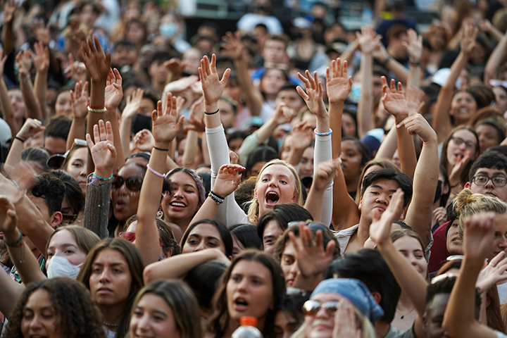 A large crowd of students hold their hands in the air as they smile and yell during a concert on the quad.