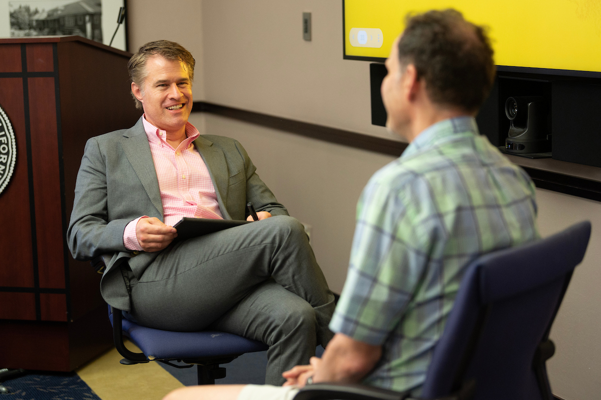 Man in gray suit smiling across from seated person in green plaid shirt in office