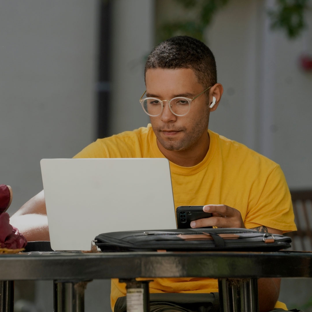 A student in a yellow shirt sits at a table on the UC Davis campus and looks at something on his laptop