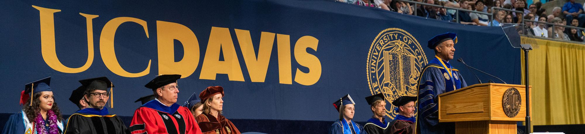 Gary May speaking at Commencement with a large UC Davis logo in the background